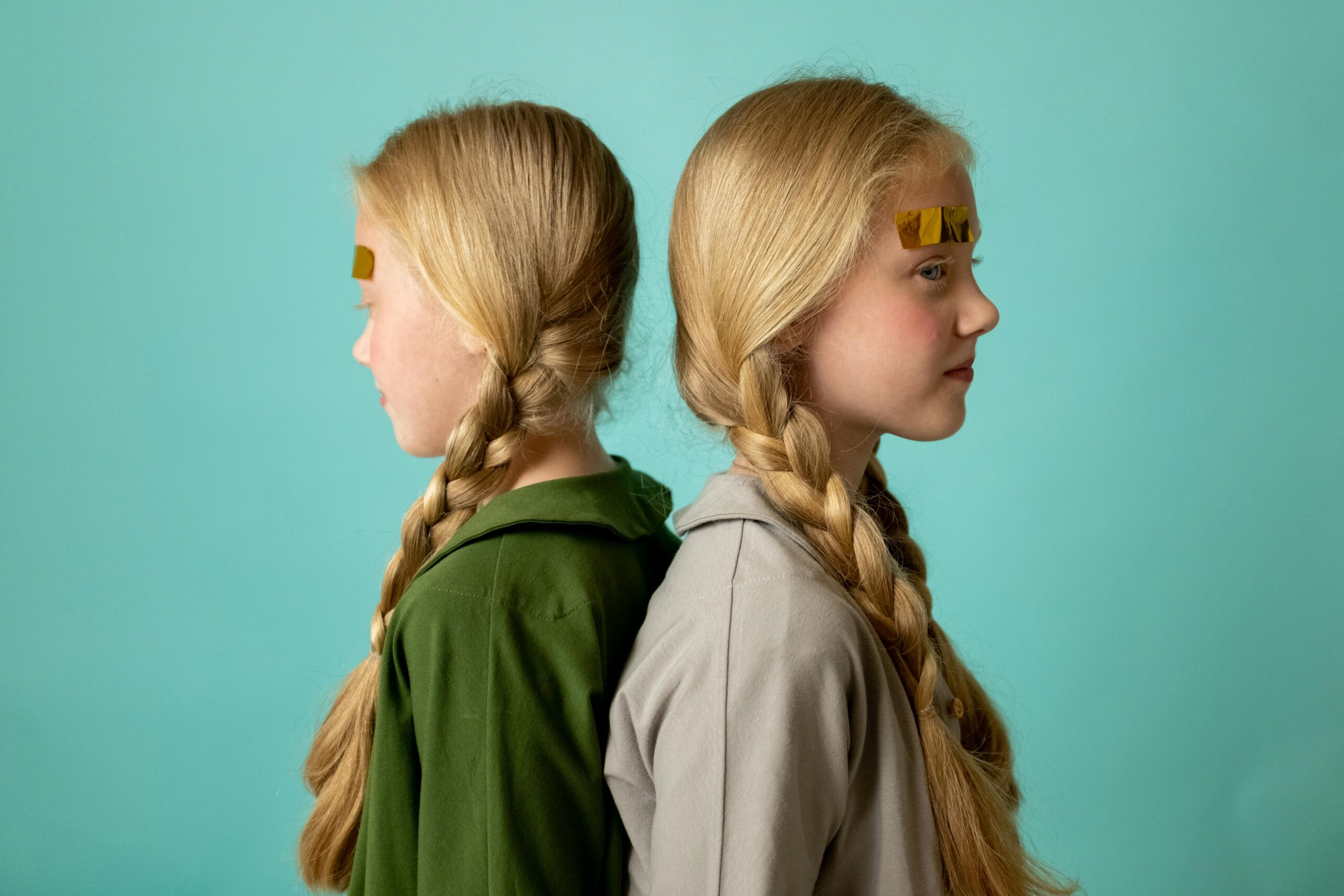 Cute twin sisters with braided hair, back to back indoors against a turquoise background.